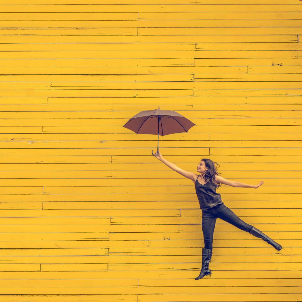 Unser Angebot: Individuell, praxiserprobt, wirksam - Institut Phoenix # Frau mit Regenschirm springt vor gelber Wand - Bild von Edu Lauton über Unsplash